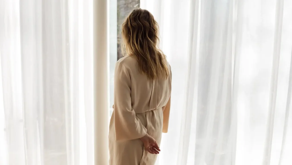 kvinde står ved foldegardiner som er hvide gardiner og kigger ud af vinduet