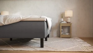 Moderne soveværelse med en Emma boxmadras, hvid dyne, natbord i lyst træ med lampe, bøger og vase. Lys geometrisk tæppe på gulvet.